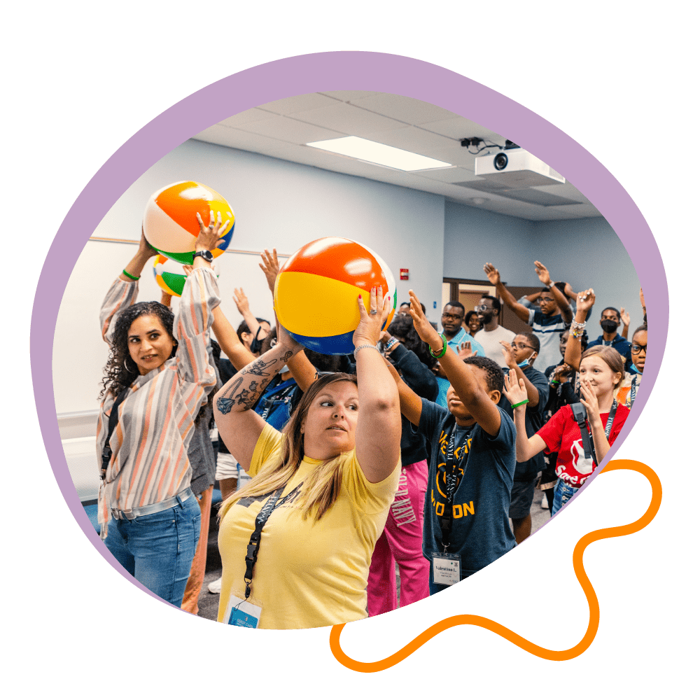 A group of educators and youth play an icebreaker game, passing beach balls overhead.